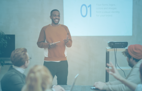 A man stands Infront of a projector screen giving a presentation to colleagues.